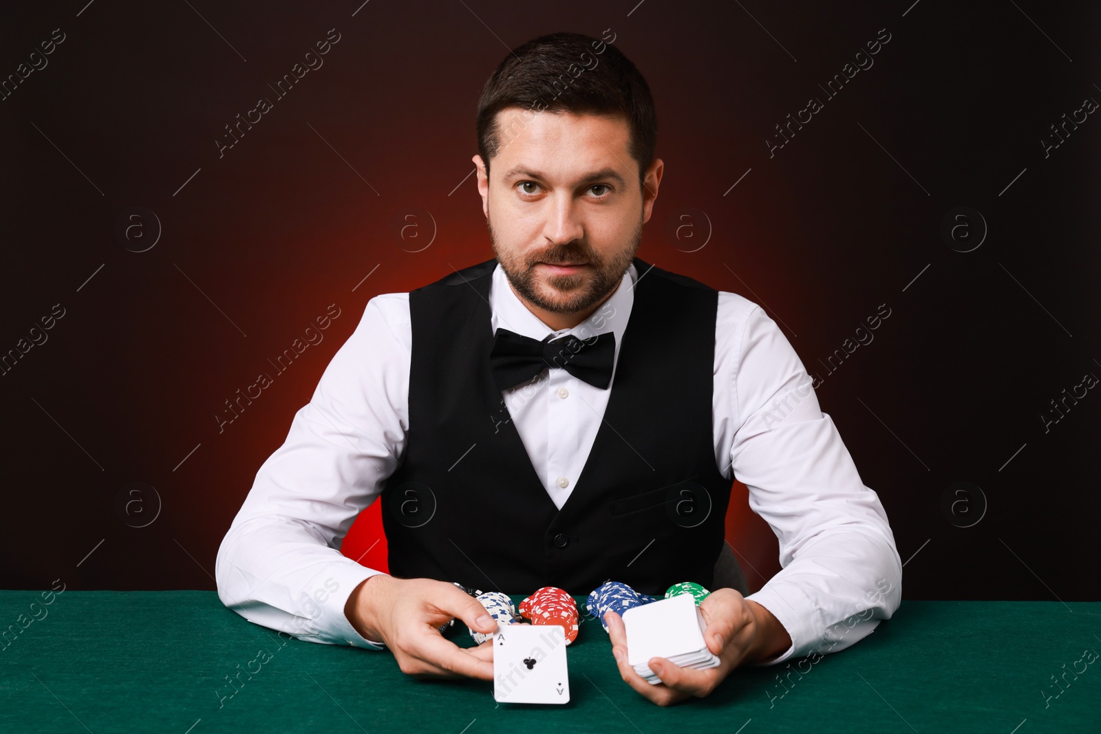 Photo of Professional croupier with playing cards at gambling table against dark red background