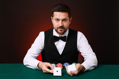 Photo of Professional croupier with playing cards at gambling table against dark red background