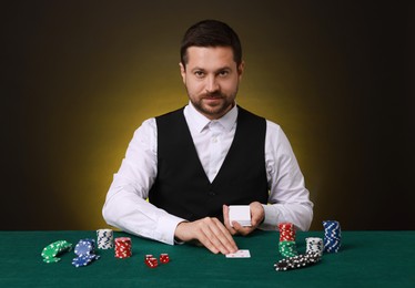 Photo of Professional croupier with playing cards at gambling table against dark yellow background