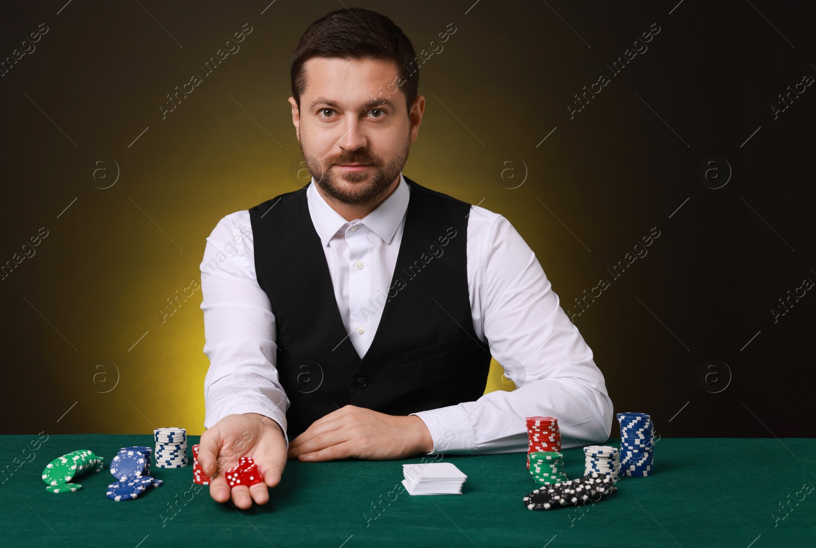 Photo of Professional croupier with playing cards at gambling table against dark yellow background