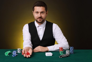 Photo of Professional croupier with playing cards at gambling table against dark yellow background