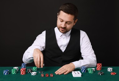 Photo of Professional croupier at gambling table with playing cards, casino chips and dice against black background