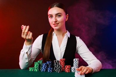 Photo of Professional croupier with casino chips and playing cards at gambling table on color background with smoke