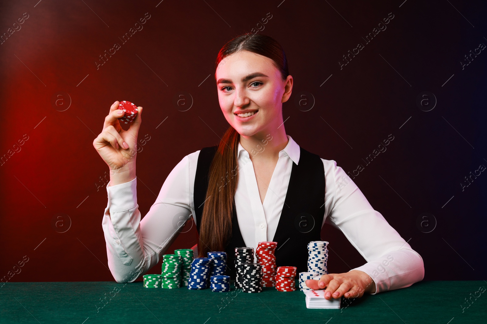 Photo of Professional croupier with casino chips and playing cards at gambling table on color background with smoke