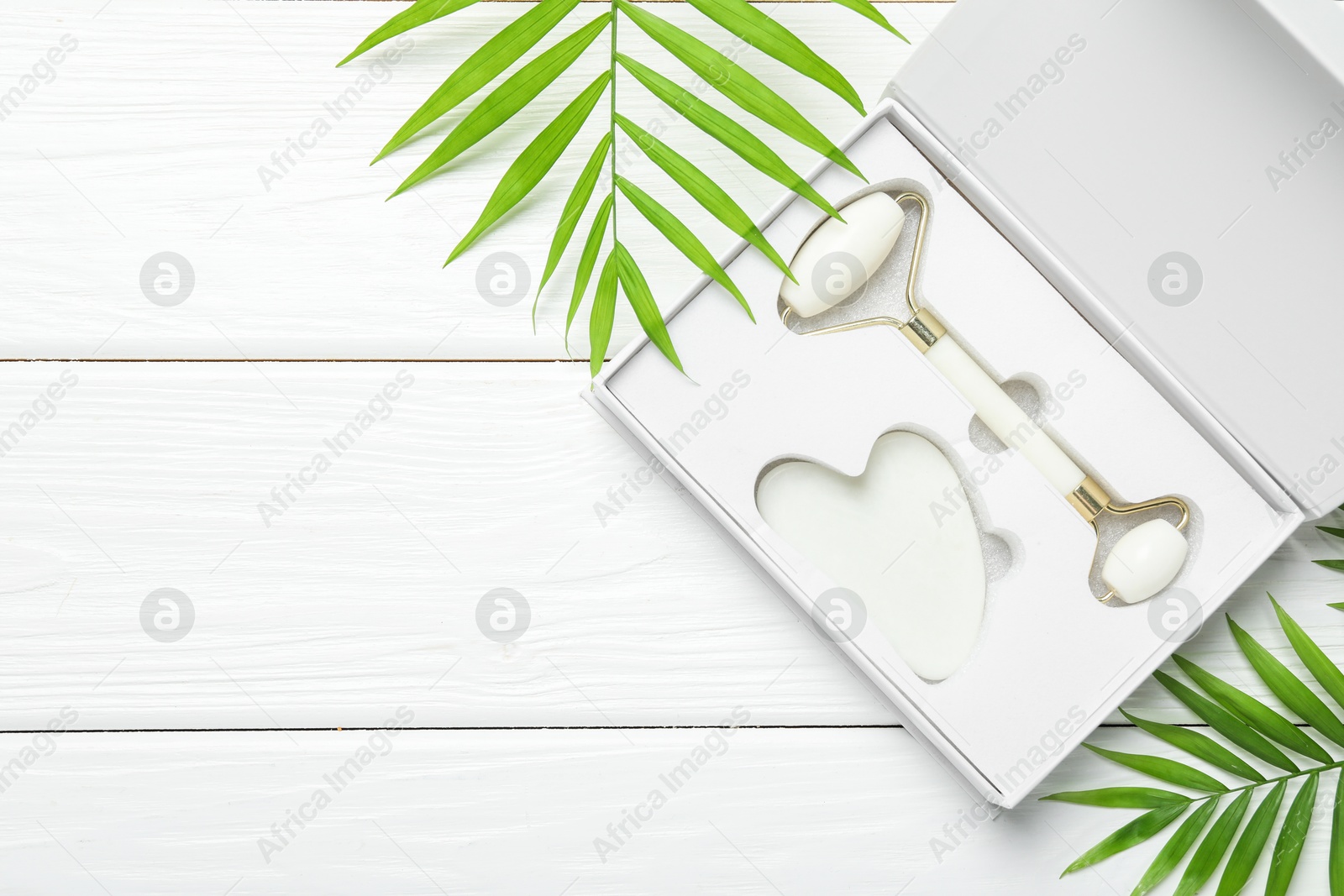 Photo of Face roller, gua sha tool in box and palm leaves on white wooden background, flat lay. Space for text