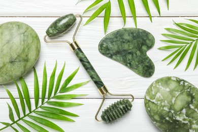 Photo of Face roller, gua sha tool, massage stones and palm leaves on white wooden background, flat lay