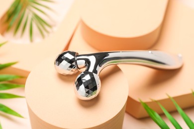 Photo of Metal face roller and palm leaves on beige background, closeup