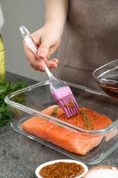 Photo of Woman marinating salmon fillet in dish at gray textured table, closeup