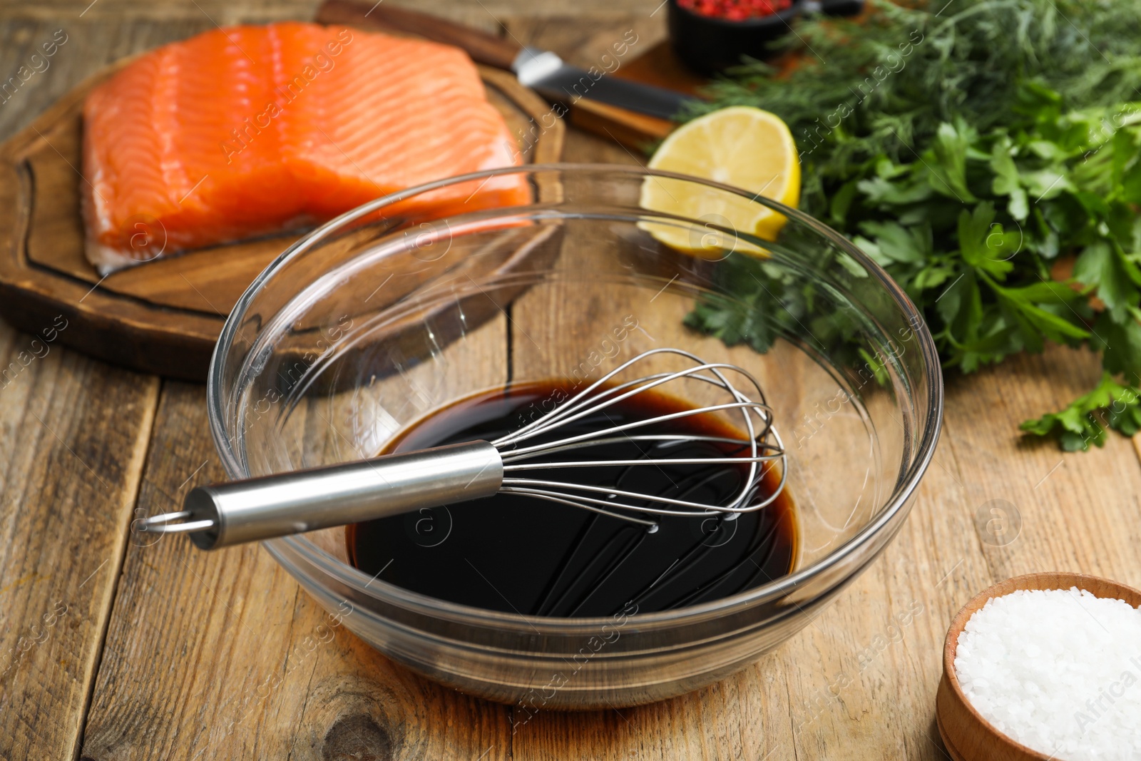 Photo of Soy sauce in bowl, whisk, salmon fillet, herbs and salt on wooden table, closeup