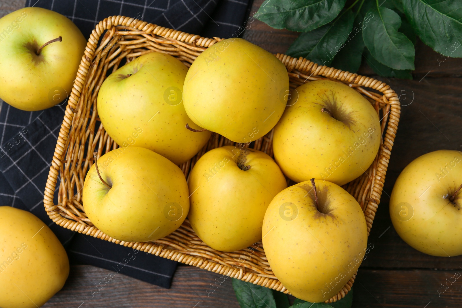 Photo of Fresh yellow apples in wicker basket and green leaves on wooden table, top view