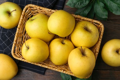 Photo of Fresh yellow apples in wicker basket and green leaves on wooden table, top view