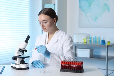 Photo of Laboratory testing. Doctor dripping blood sample onto glass slide while working with microscope at table indoors