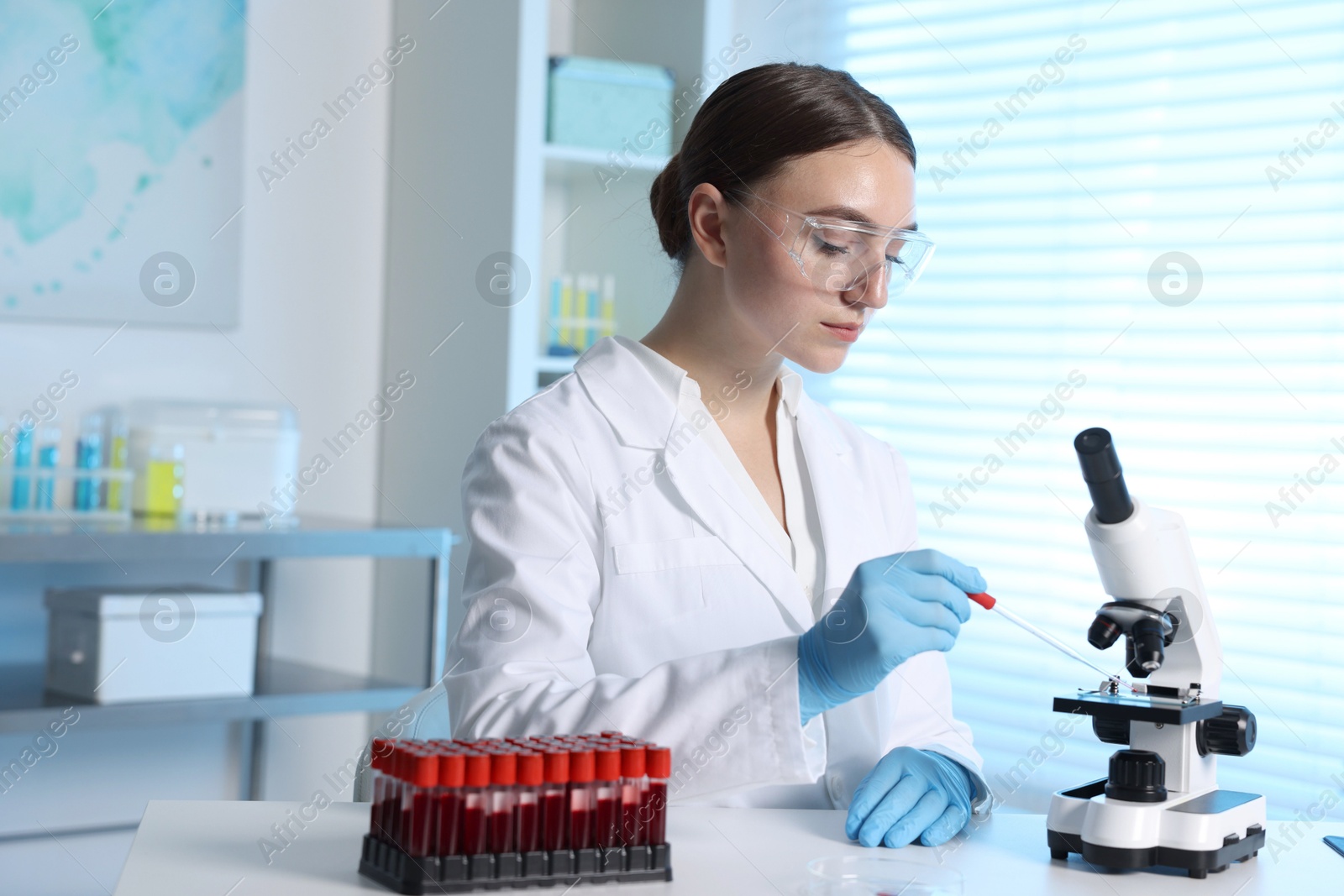 Photo of Laboratory testing. Doctor dripping blood sample onto glass slide while working with microscope at table indoors