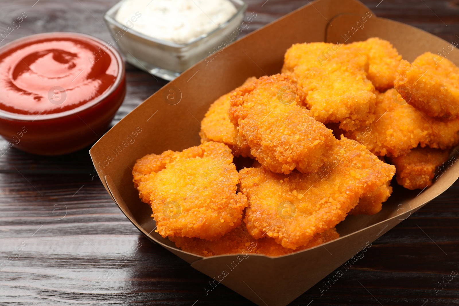 Photo of Tasty chicken nuggets in paper container and sauces on wooden table, closeup