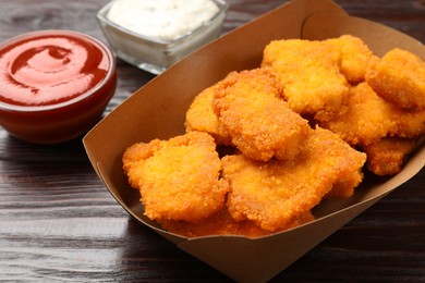 Photo of Tasty chicken nuggets in paper container and sauces on wooden table, closeup