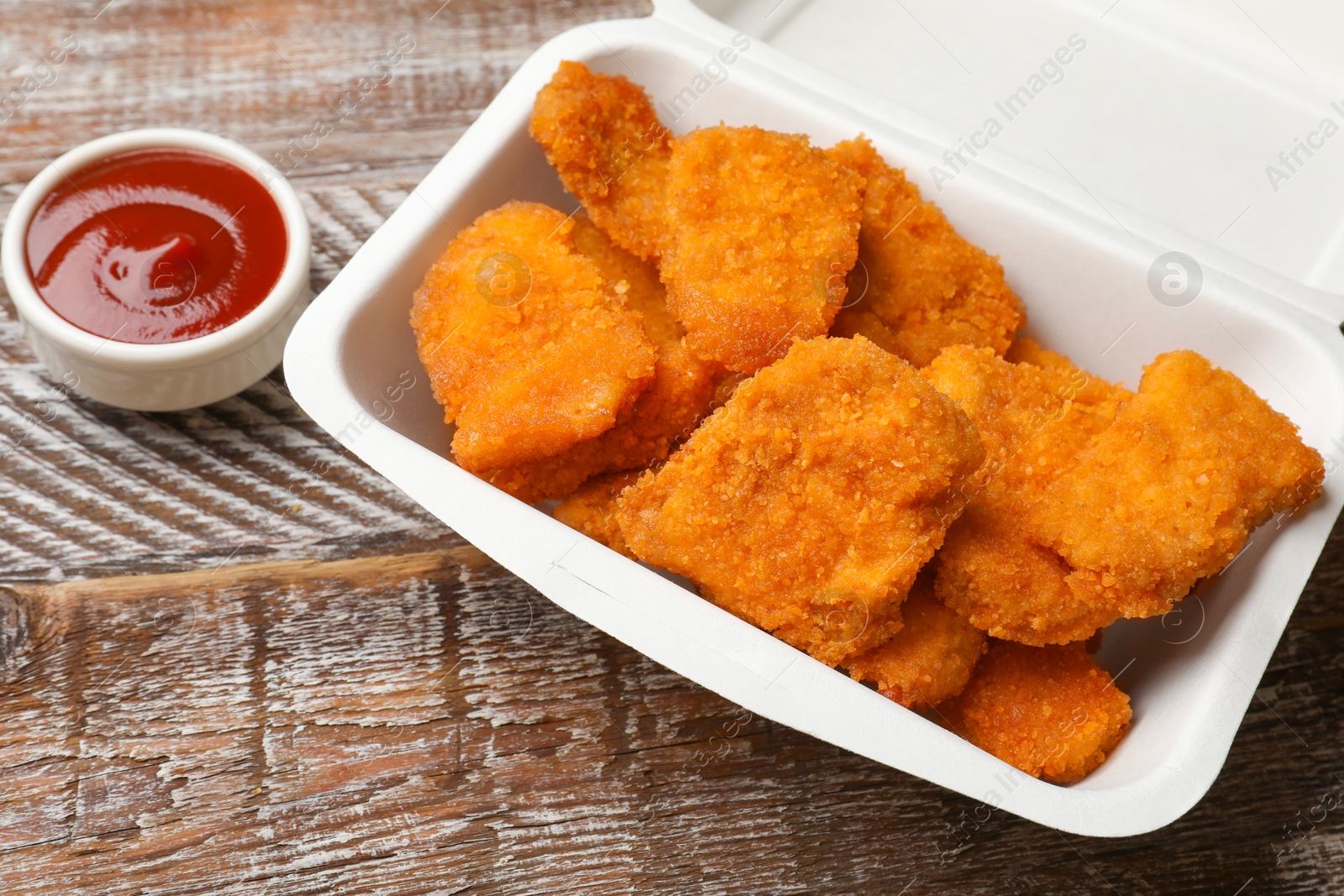 Photo of Delicious chicken nuggets in takeaway container and ketchup on wooden table, closeup