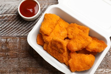 Photo of Delicious chicken nuggets in takeaway container and ketchup on wooden table, above view