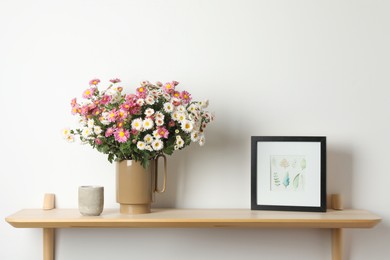 Photo of Vase with beautiful flowers and other decor on wooden shelf near white wall