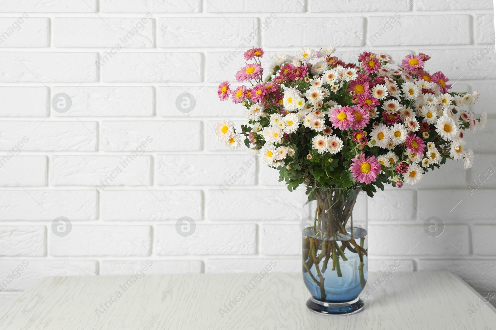 Photo of Vase with beautiful flowers on wooden table near white brick wall, space for text