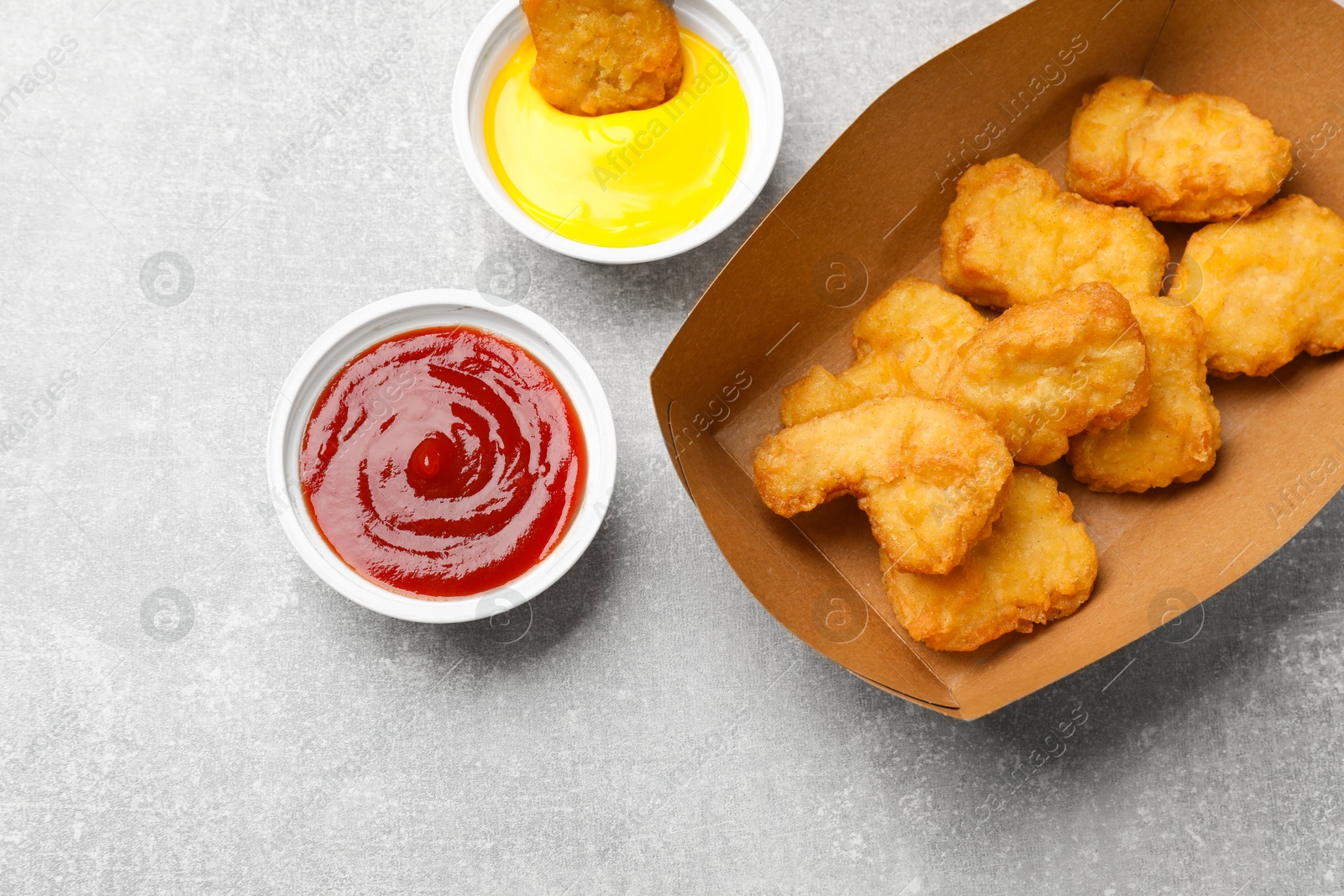 Photo of Delicious chicken nuggets in carton box and sauces on gray textured table, flat lay