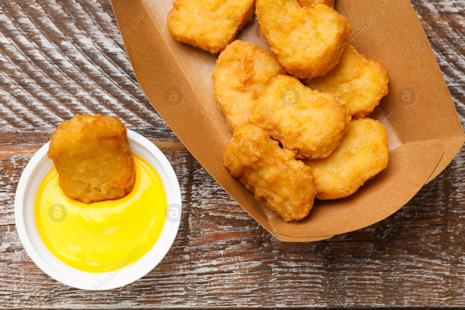 Photo of Delicious chicken nuggets in carton box and sauce on wooden table, flat lay