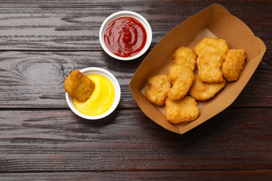 Photo of Delicious chicken nuggets in carton box and sauces on wooden table, flat lay