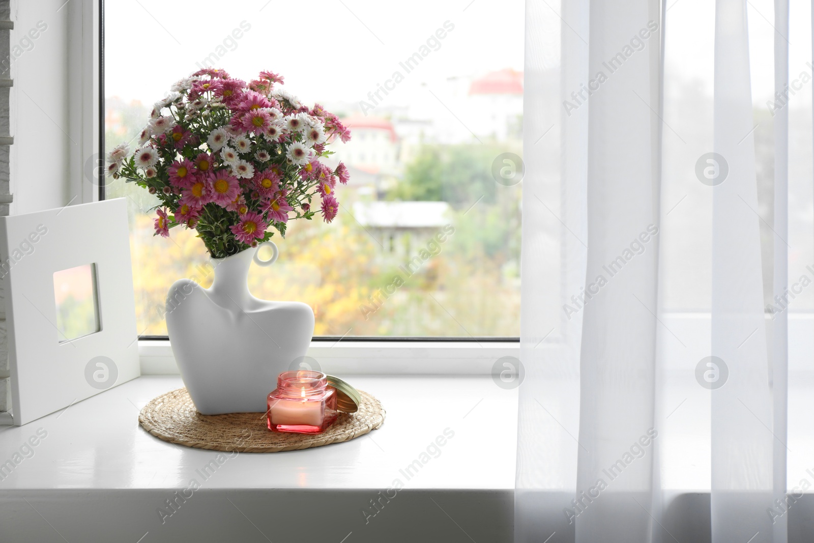 Photo of Stylish vase with beautiful flowers and burning candle near window at home