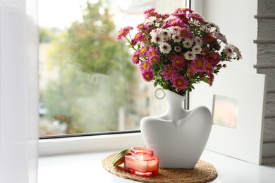 Photo of Stylish vase with beautiful flowers and burning candle near window at home