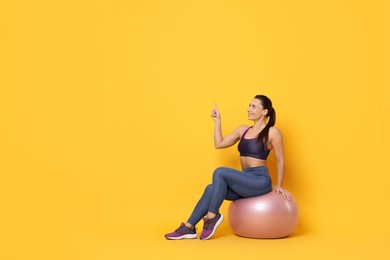 Photo of Smiling woman in sportswear sitting on fitness ball and pointing upwards against yellow background. Space for text