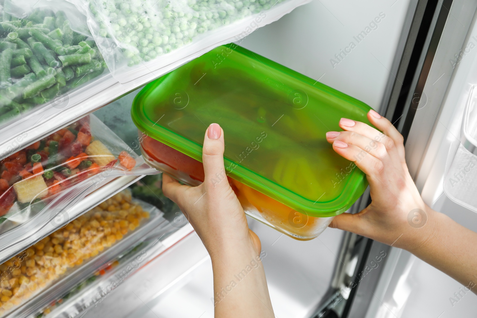 Photo of Woman taking glass container with frozen cut bell pepper from refrigerator, closeup