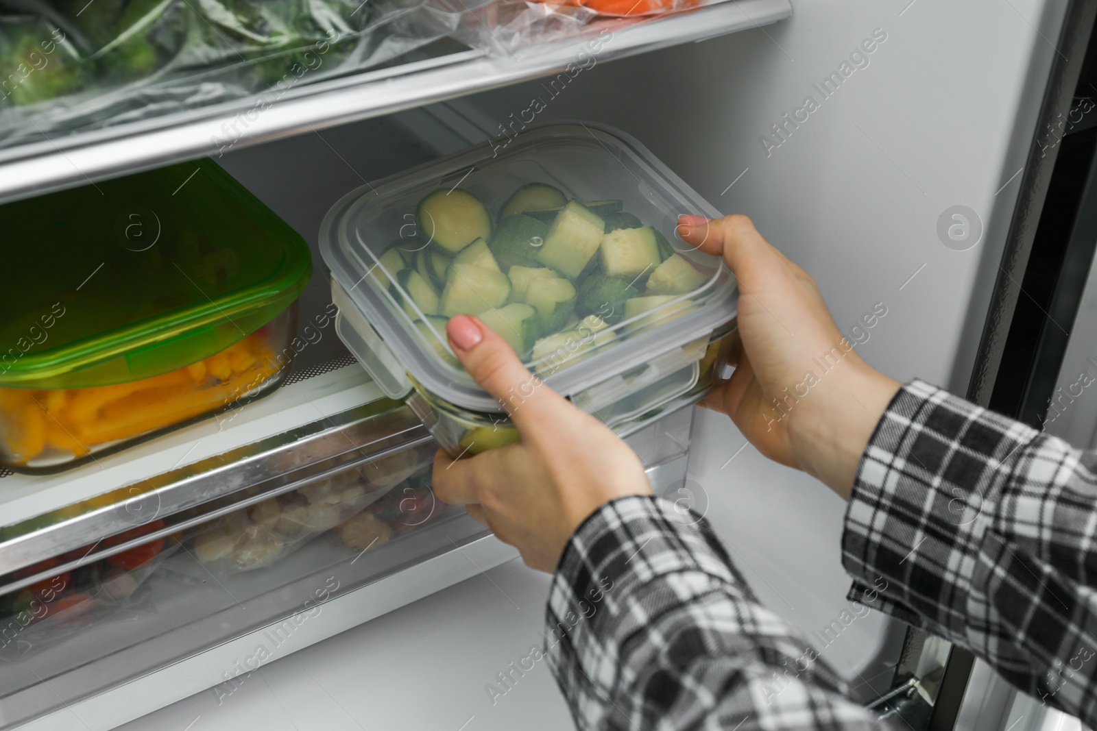 Photo of Woman taking glass container with frozen chopped zucchini from refrigerator, closeup