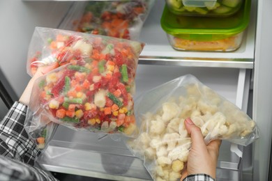 Photo of Woman taking plastic bags with mix of frozen vegetables and cauliflower, from refrigerator, closeup