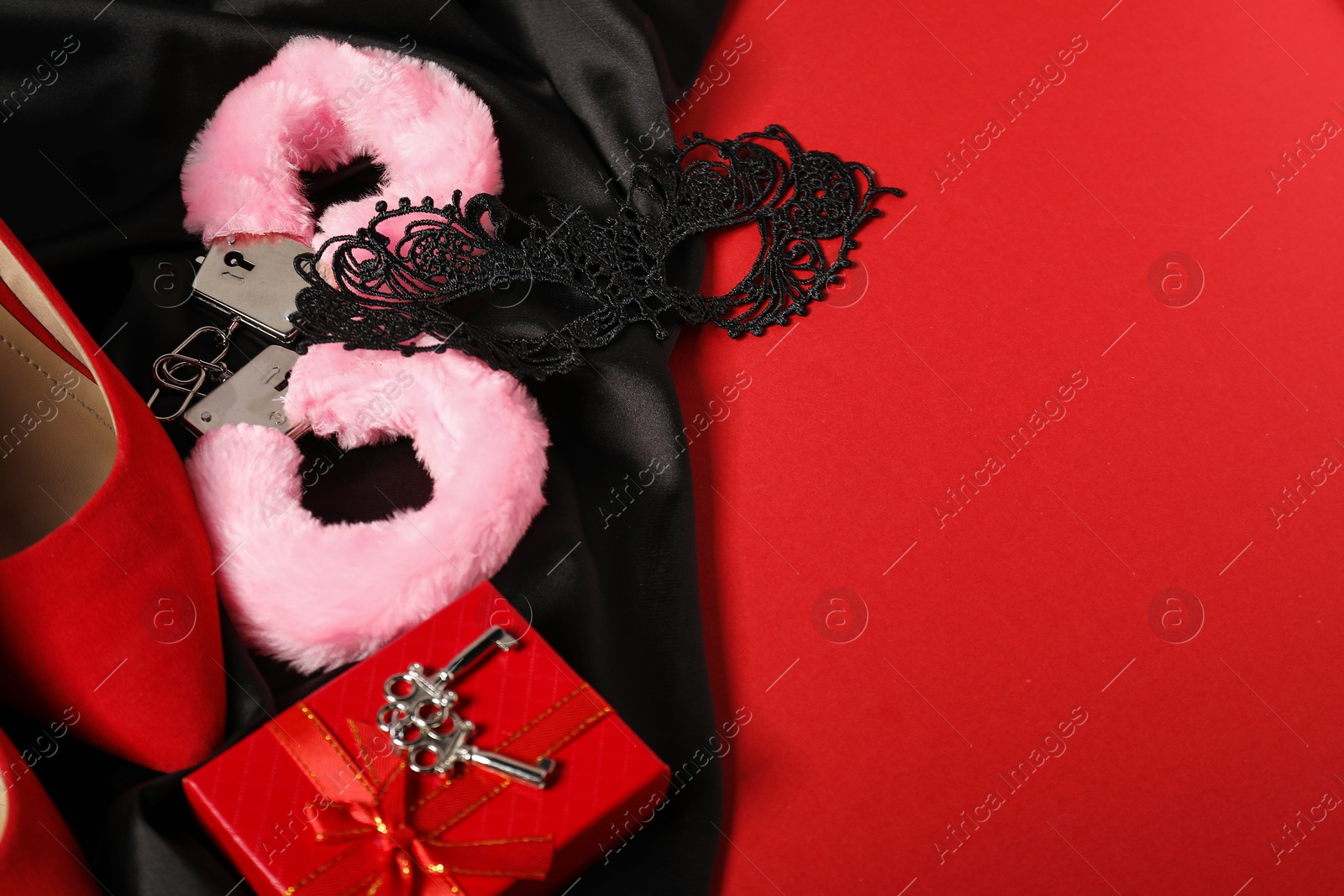 Photo of Pink fluffy handcuffs, keys, lace mask, shoes and black fabric on red background. Space for text