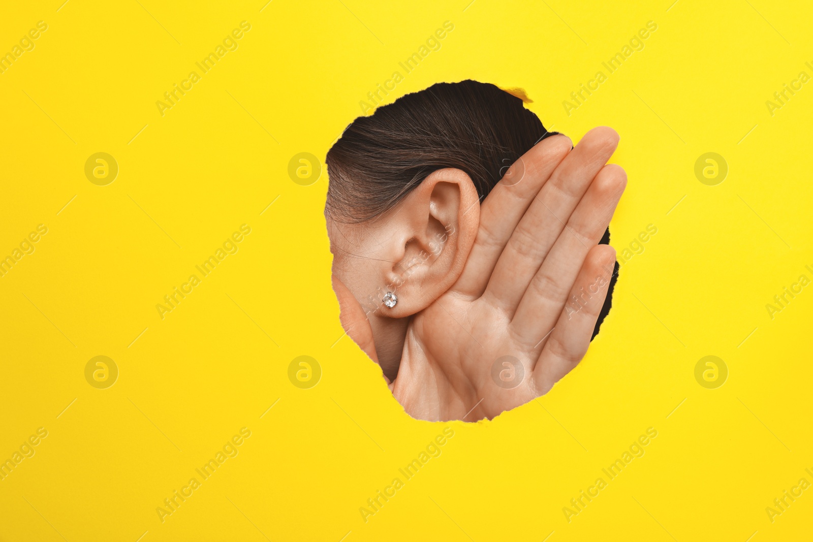 Photo of Woman showing hand to ear gesture through hole in yellow paper, closeup