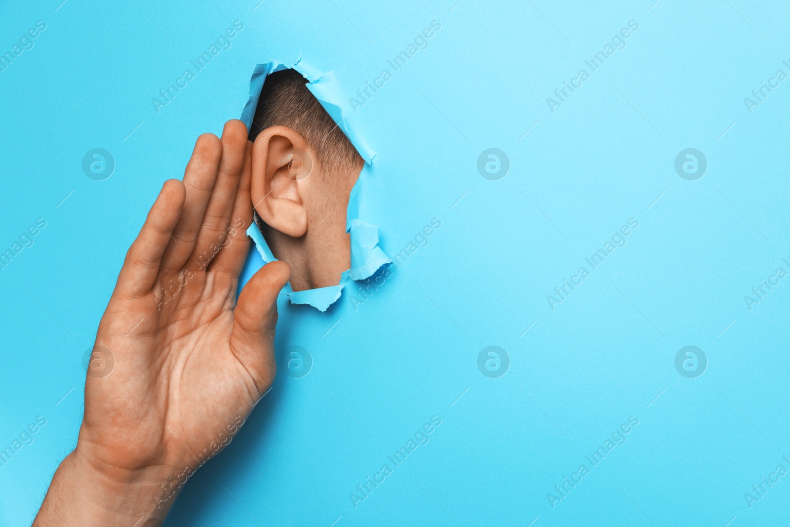 Photo of Man showing hand to ear gesture through hole in light blue paper, closeup. Space for text