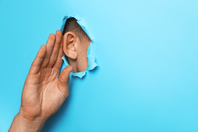 Man showing hand to ear gesture through hole in light blue paper, closeup. Space for text