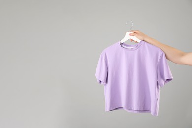 Photo of Woman holding hanger with violet t-shirt on grey background, closeup. Space for text