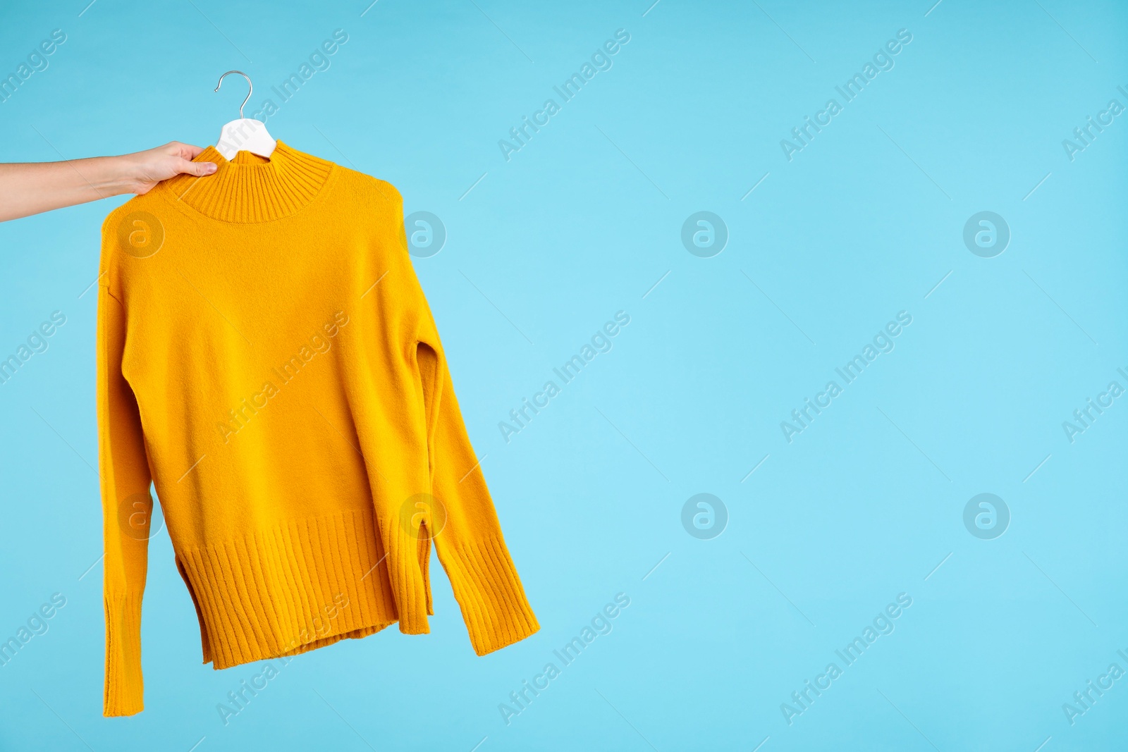 Photo of Woman holding hanger with warm sweater on light blue background, closeup. Space for text