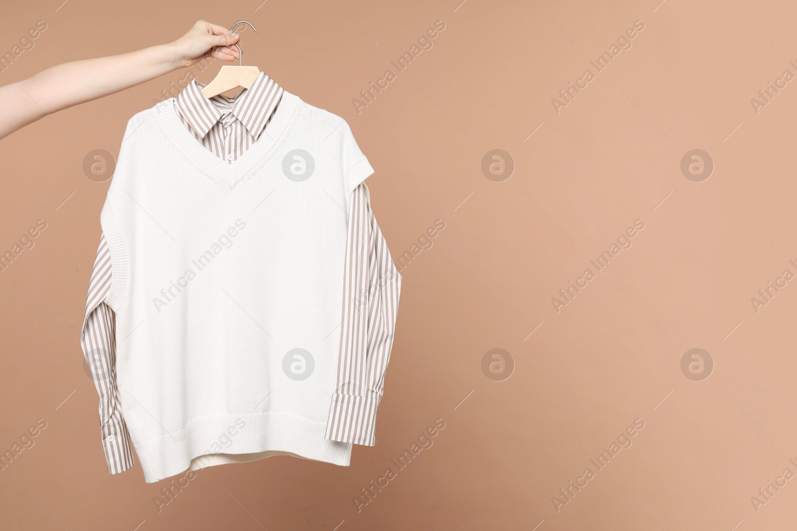 Photo of Woman holding hanger with shirt and sweater vest on beige background, closeup. Space for text