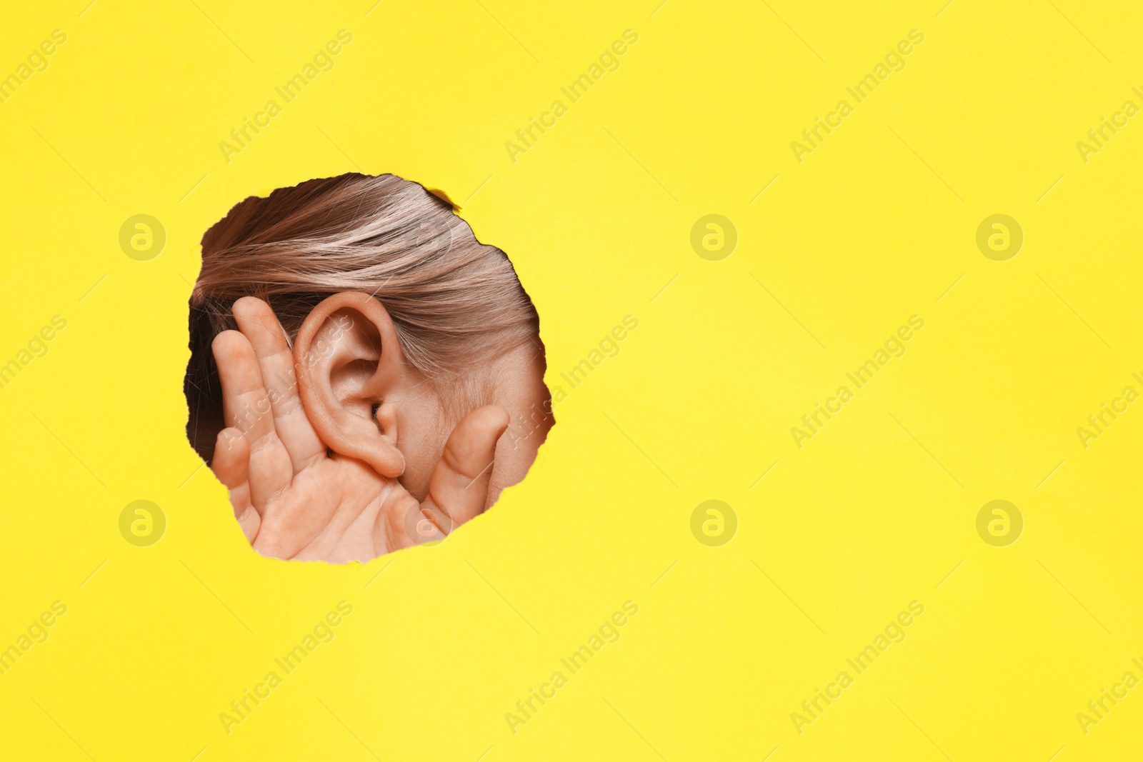 Photo of Little girl showing hand to ear gesture through hole in yellow paper, closeup. Space for text