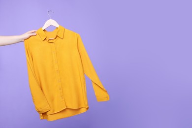 Photo of Woman holding hanger with orange shirt on light purple background, closeup. Space for text