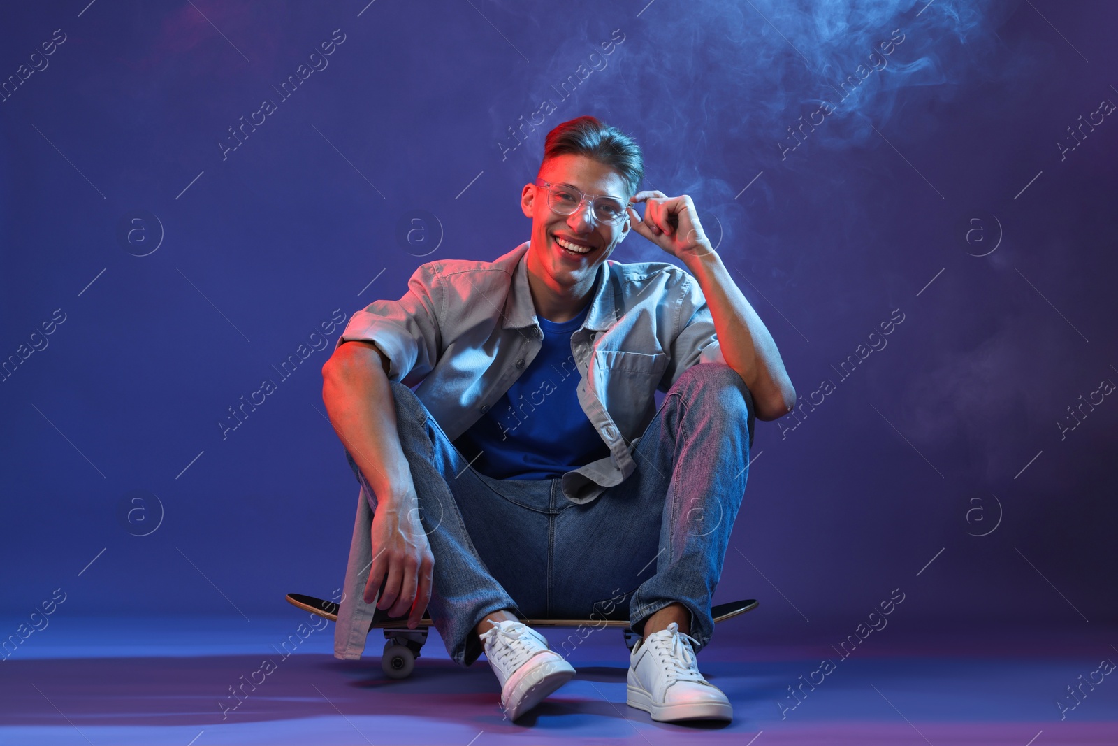 Photo of Happy man sitting on skateboard in smoke and color lights against blue background