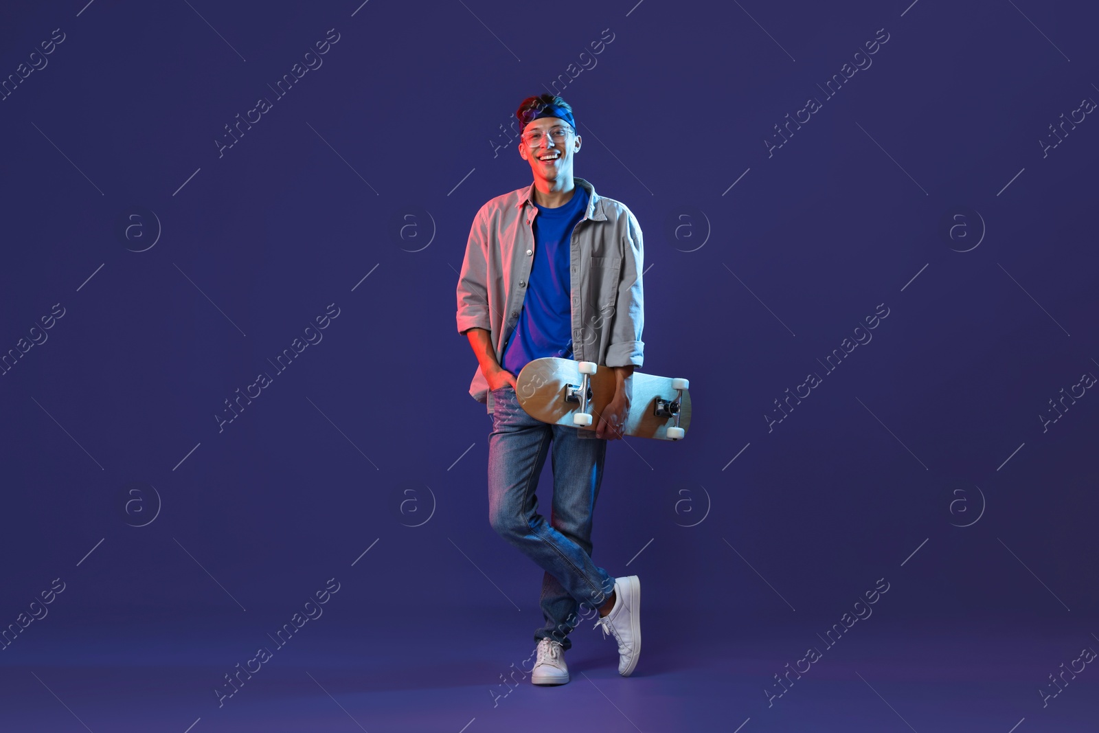 Photo of Happy man with skateboard in color lights against blue background