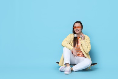 Smiling woman sitting on skateboard against light blue background. Space for text