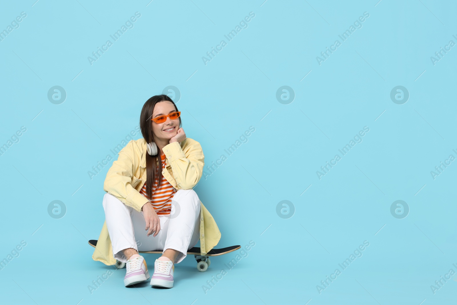 Photo of Smiling woman sitting on skateboard against light blue background. Space for text