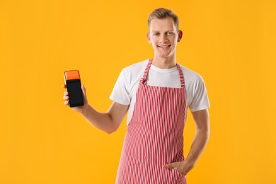 Happy young man in apron with payment terminal on yellow background