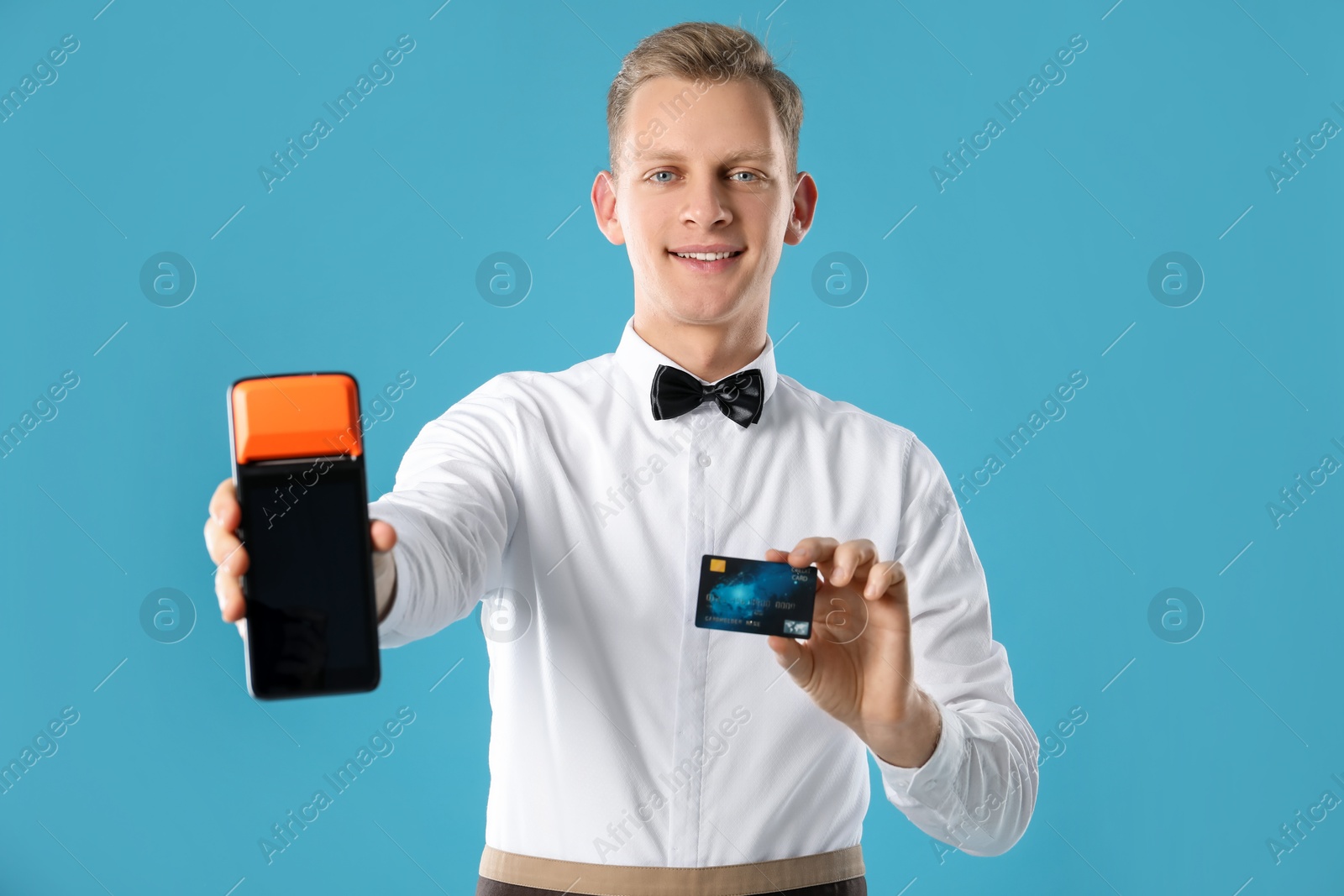 Photo of Happy waiter with payment terminal and debit card on light blue background