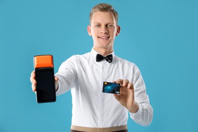 Happy waiter with payment terminal and debit card on light blue background