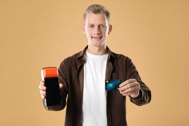 Happy young man with payment terminal and debit card on beige background