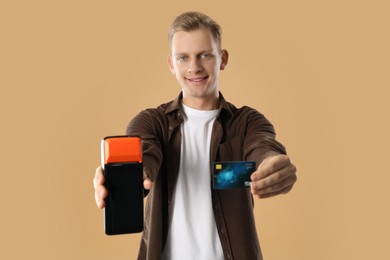 Photo of Happy young man with payment terminal and debit card on beige background